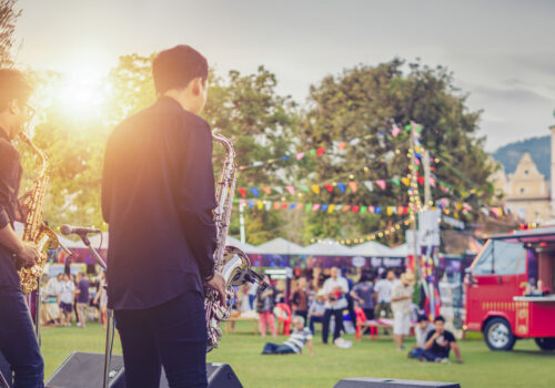 Zwei Musiker mit Saxophon auf einer Bühne bei einem sommerlichen Stadtfest mit Foodtruck, Lichterketten und Publikum im Hintergrund – stimmungsvolles Beispiel dafür, wie Städte erfolgreich City Events planen und beleben.