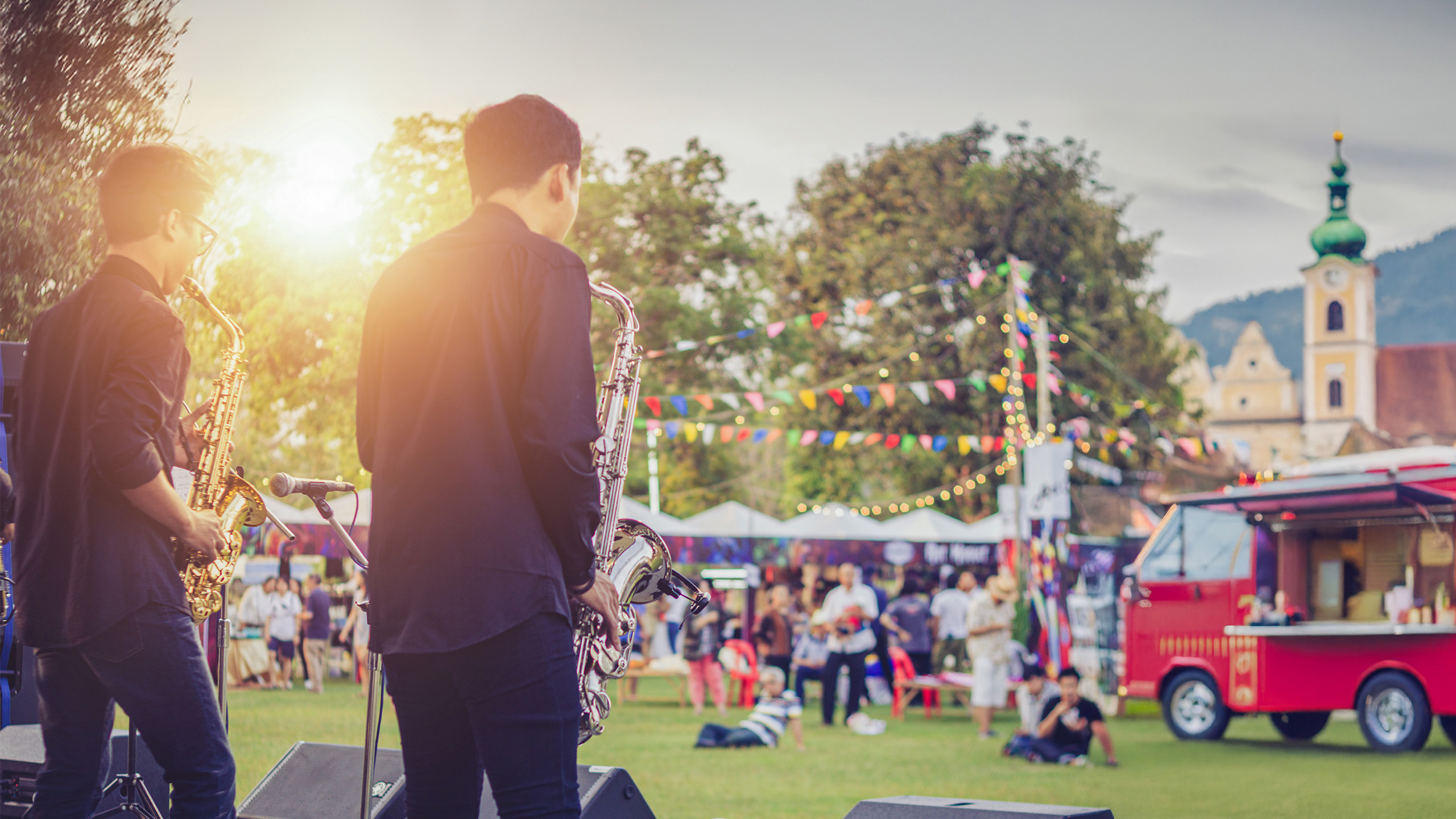 Zwei Musiker mit Saxophon auf einer Bühne bei einem sommerlichen Stadtfest mit Foodtruck, Lichterketten und Publikum im Hintergrund – stimmungsvolles Beispiel dafür, wie Städte erfolgreich City Events planen und beleben.