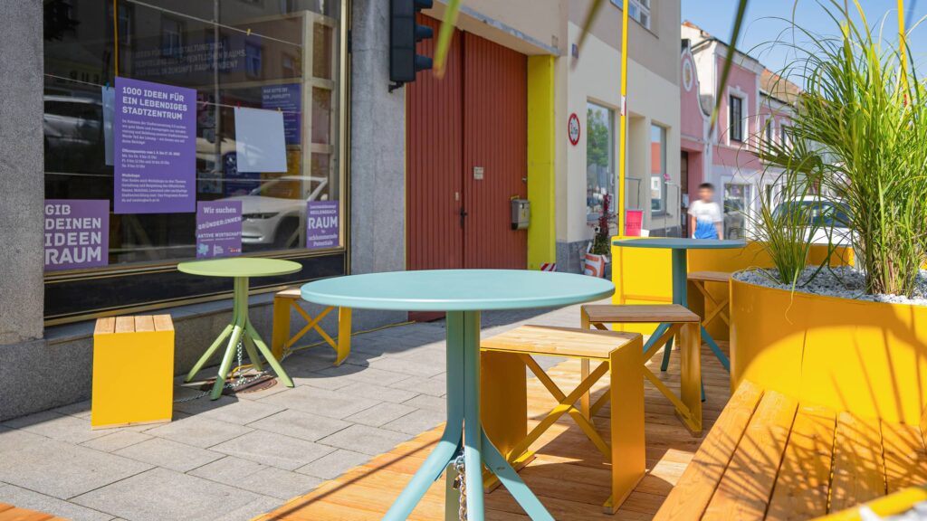 Detailaufnahme eines Parklets mit bunten Sitzmöbeln und Pflanzen vor einem leerstehenden Geschäft – Zwischennutzung zur Förderung von Aufenthaltsqualität und Leerstandsaktivierung.