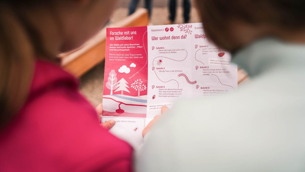 Zwei Kinder betrachten gemeinsam ein Informationsblatt mit dem Titel "Forsche mit uns im Waldlabor".