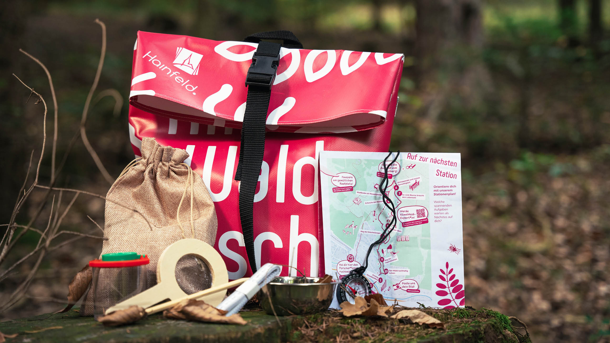 Forscherrucksack mit Wald-Experimentierutensilien wie Lupe, Becherlupe, Kompass und Karte, ausgebreitet im Wald.