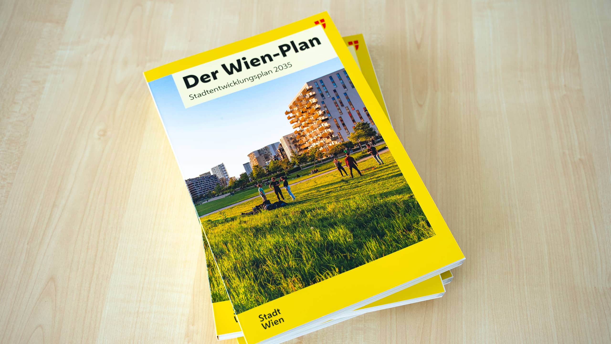 Gedruckte Broschüre des Wien-Plans – Stadtentwicklungsplan 2035 – mit gelbem Einband, einem Foto von Menschen auf einer Wiese vor Wohngebäuden und dem Logo der Stadt Wien, liegend auf einem hellen Holztisch