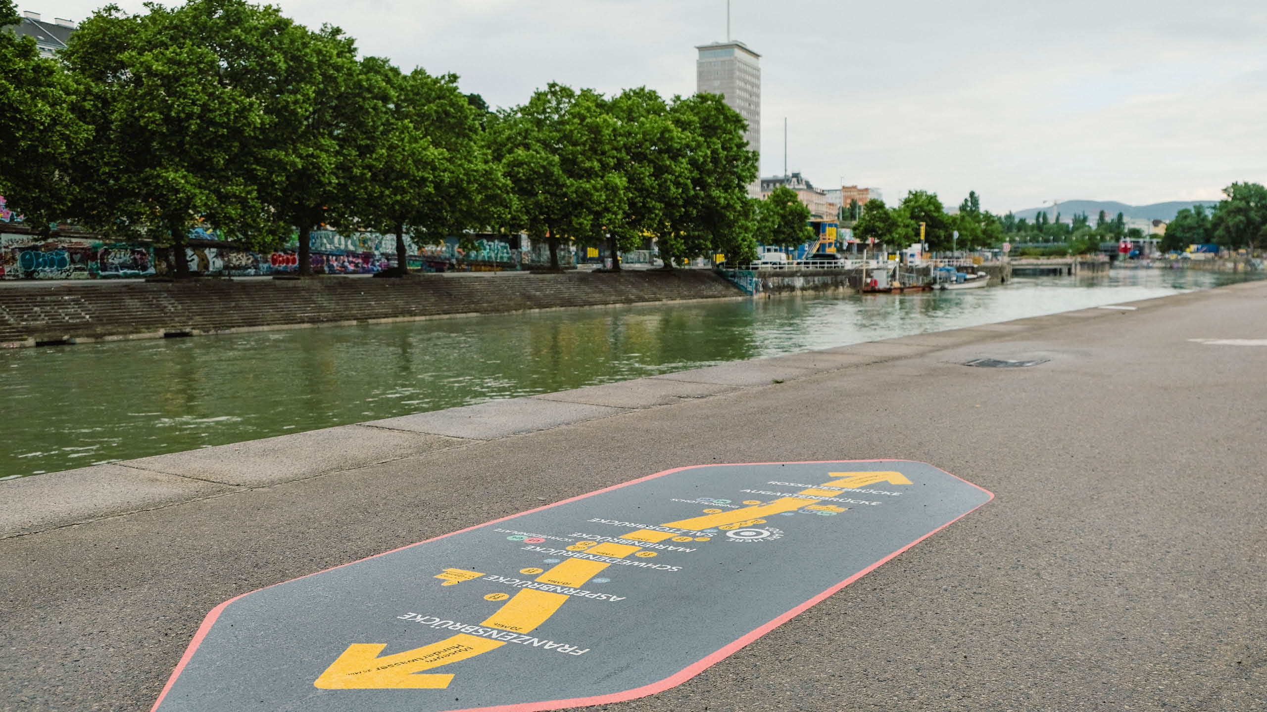 Blick entlang des Donaukanals in Wien mit neu gestalteter Bodenmarkierung, die auf Brücken und Gehzeiten hinweist. Im Hintergrund Bäume und Graffiti.