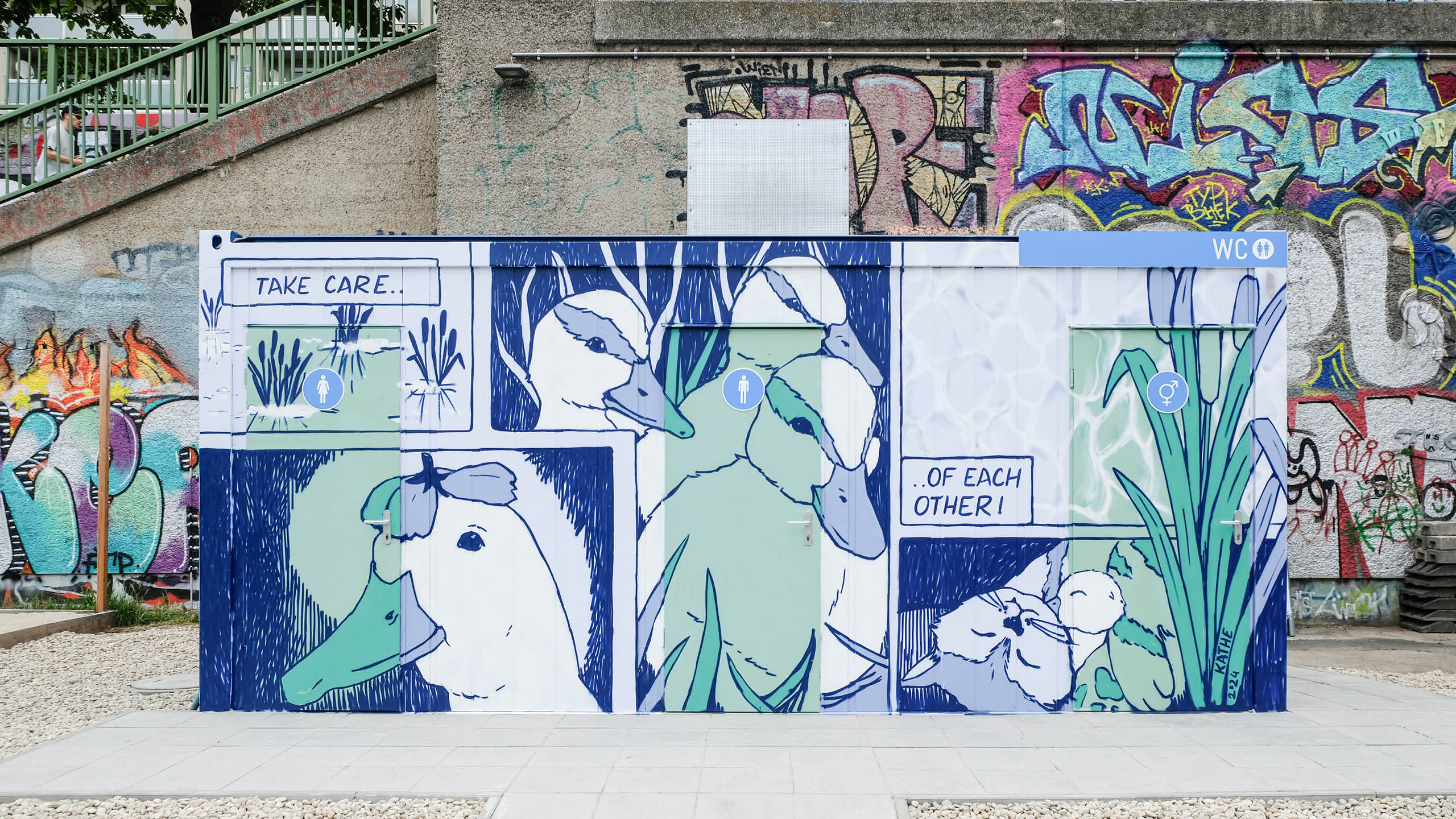 Komplettansicht der bemalten WC-Container am Donaukanal mit Enten-Cartoons und der Aufschrift „Take care… of each other!“, künstlerisch gestaltet von Käthe Löffelmann.