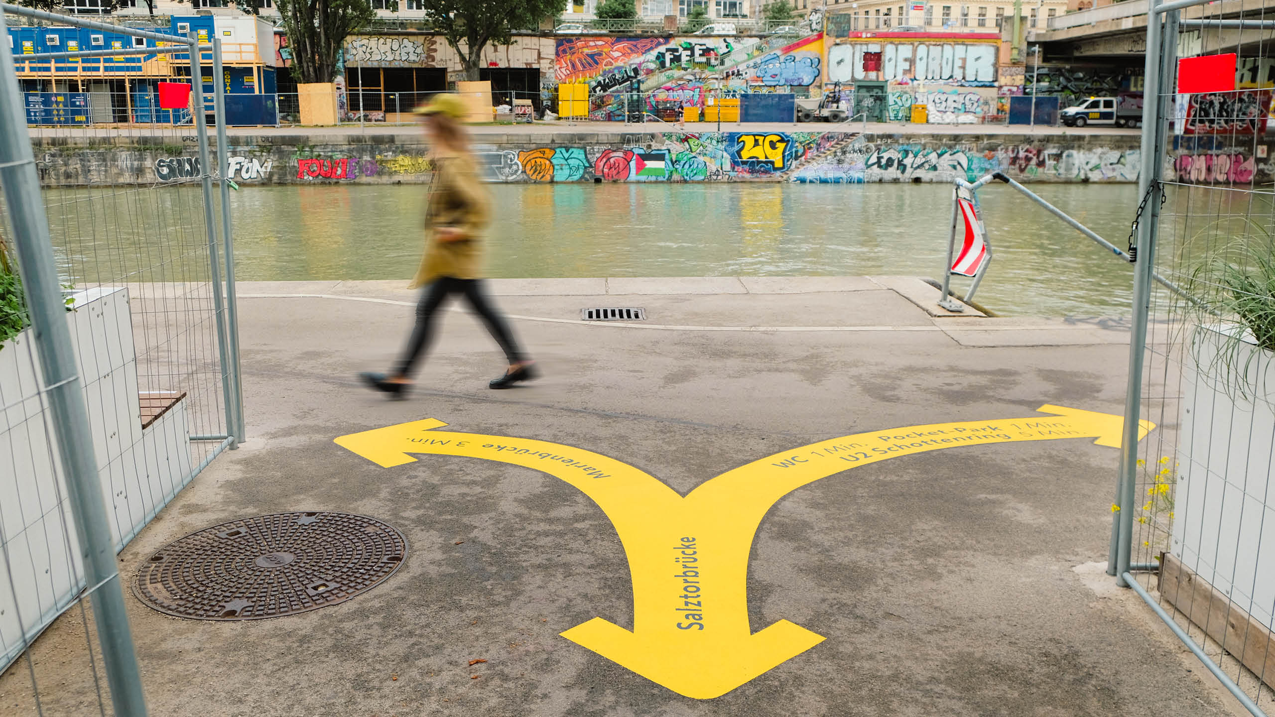 Gabelung der gelben Bodenmarkierung am Donaukanal, zeigt Wege zur Marienbrücke und zu WC/Schottenring, während eine Person vorbeigeht.