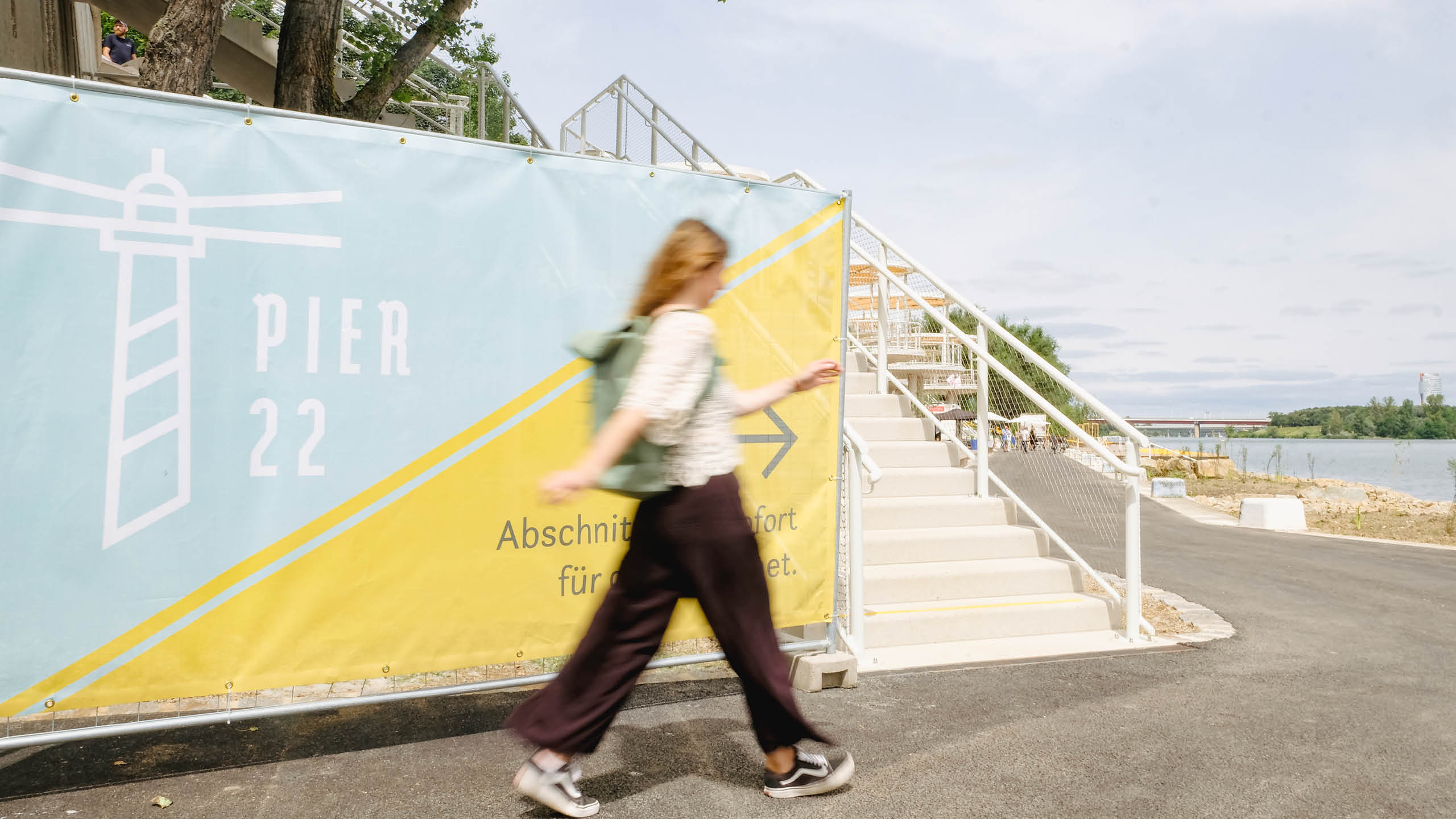 Besucher*in geht an einem bunten Banner mit PIER 22 Logo auf der Donauinsel vorbei – Sommerliches Badeareal mit konsumfreien Zonen in Wien.