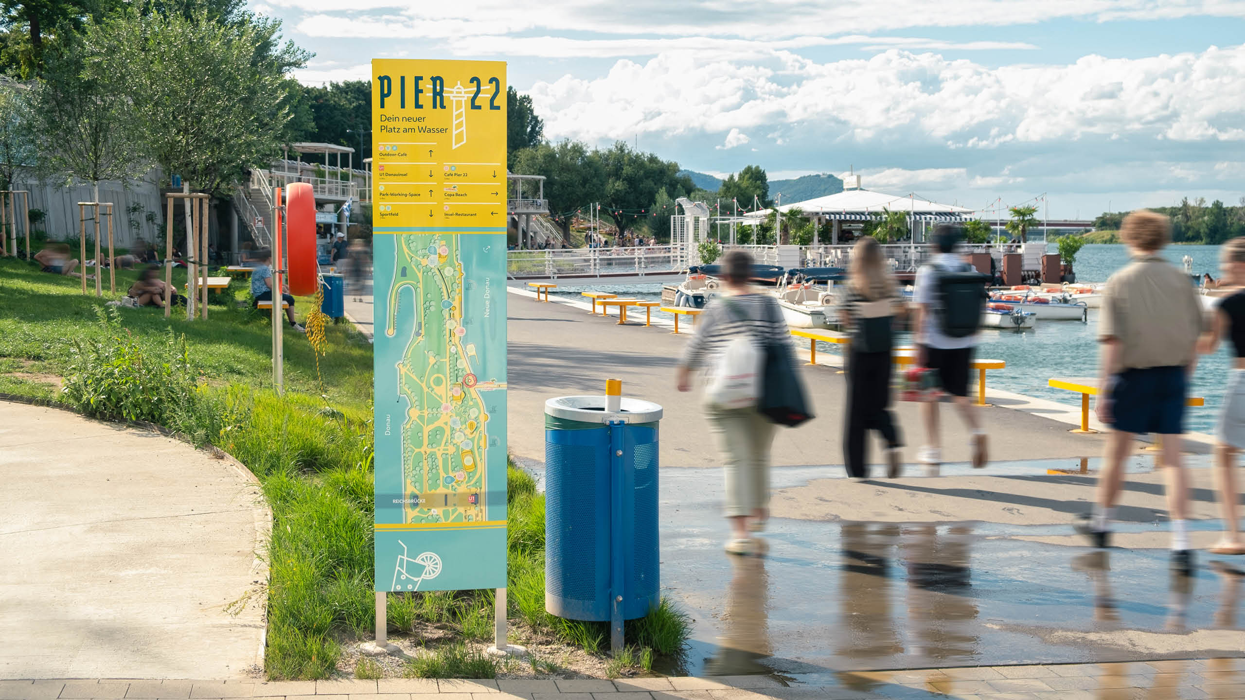 Menschen flanieren entlang der Uferpromenade auf der Donauinsel beim Pier 22. Im Vordergrund eine gelbe Infostele mit Lageplan und Richtungsanzeige. Im Hintergrund der CopaBeach und die Neue Donau.