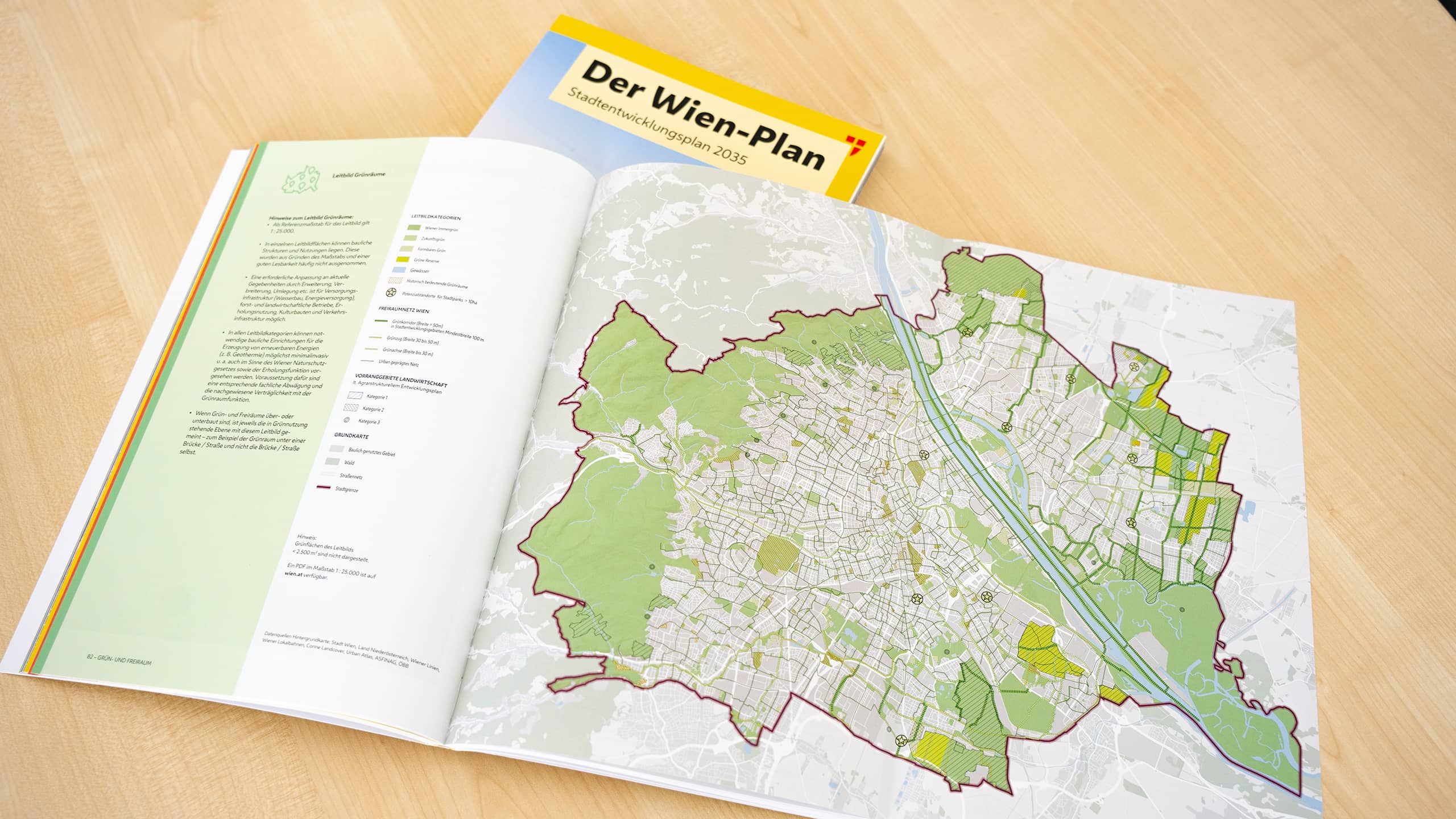 Aufgeschlagene Doppelseite des Wien-Plans 2035 auf einem Holztisch, mit einer farbigen Übersichtskarte der Stadt Wien inklusive Grünflächen und Entwicklungszonen sowie einer Legende auf der linken Seite; im Hintergrund liegt das Titelheft geschlossen