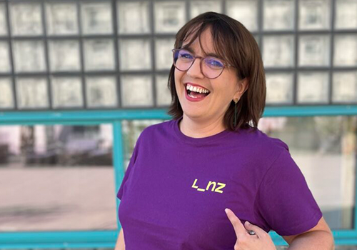 Porträt einer lachenden Silvia Hackl mit Brille im violetten T-Shirt mit gelbem „Linz“-Logo