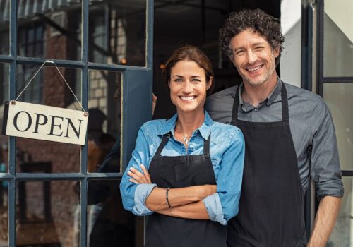 Zwei Gründer:innen vor ihrem neu eröffneten Geschäft, lächelnd im Eingangsbereich mit „Open“-Schild – Symbolbild für erfolgreiche Ansiedlung durch gezieltes Standortmarketing und eine überzeugende Standortwebsite.