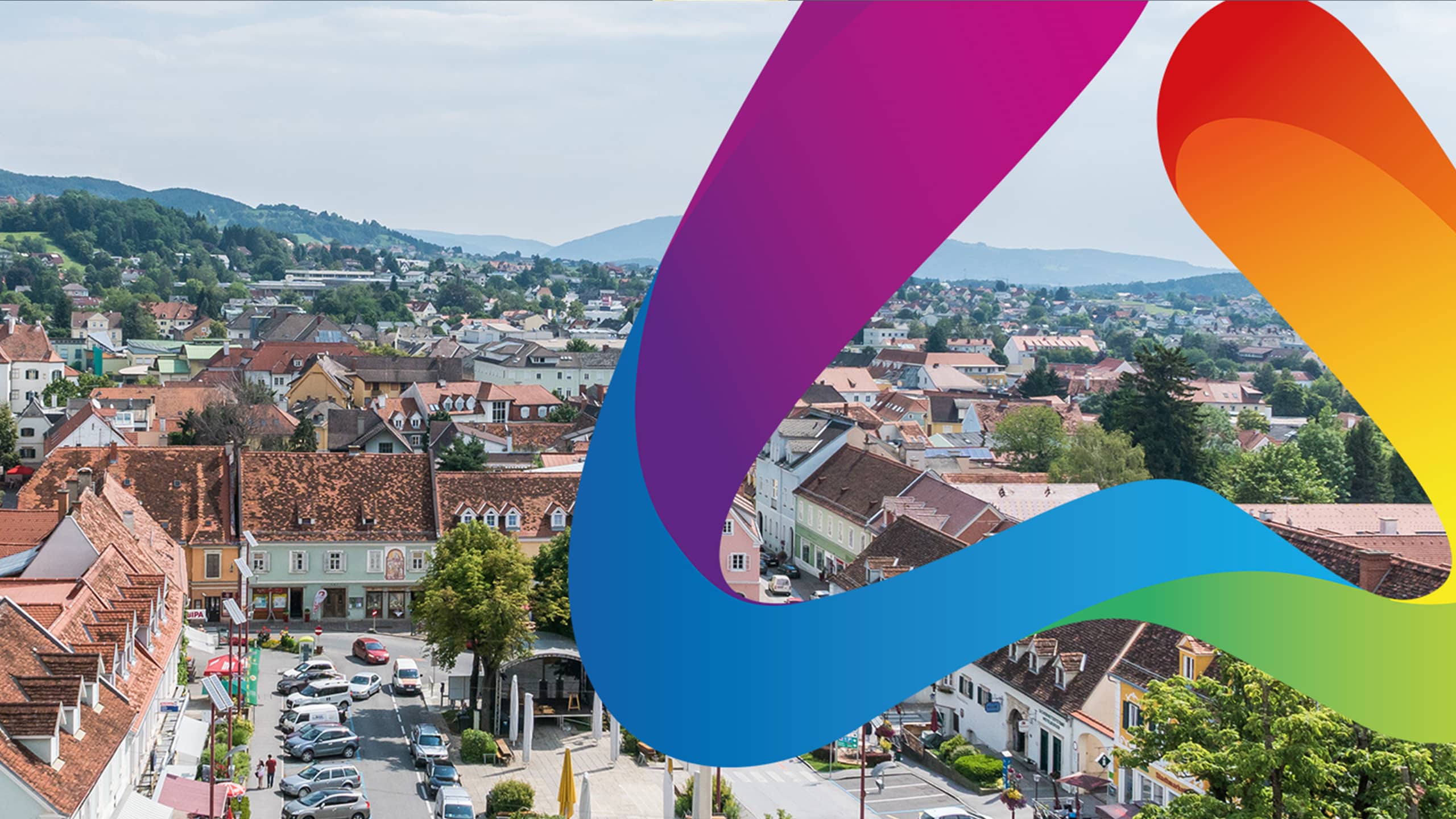 Blick über die Stadt Weiz mit historischer Dachlandschaft, Innenstadt und umliegender Hügellandschaft – im Vordergrund modernes Farbband als Branding-Element der Stadtmarke Weiz.