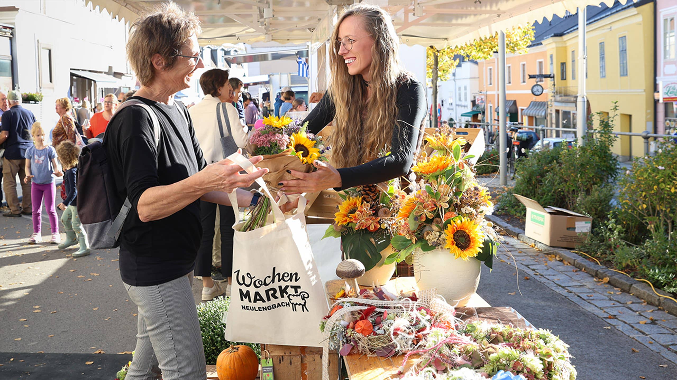 Am Wochenmarkt Neulengbach überreicht eine junge Marktfrau mit langen Haaren bunte Blumensträuße an eine Kundin. Im Hintergrund flanieren Besucher*innen durch die Marktstände in der herbstlichen Innenstadt.