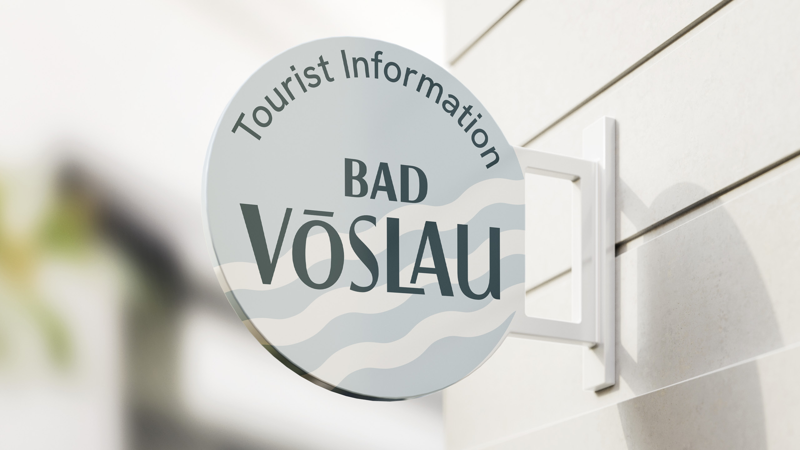 bad_voeslau_typografie_vielfalt Rundes Schild mit der Aufschrift „Tourist Information Bad Vöslau“ an einer hellen Hauswand, Gestaltung in sanften Wellenformen.